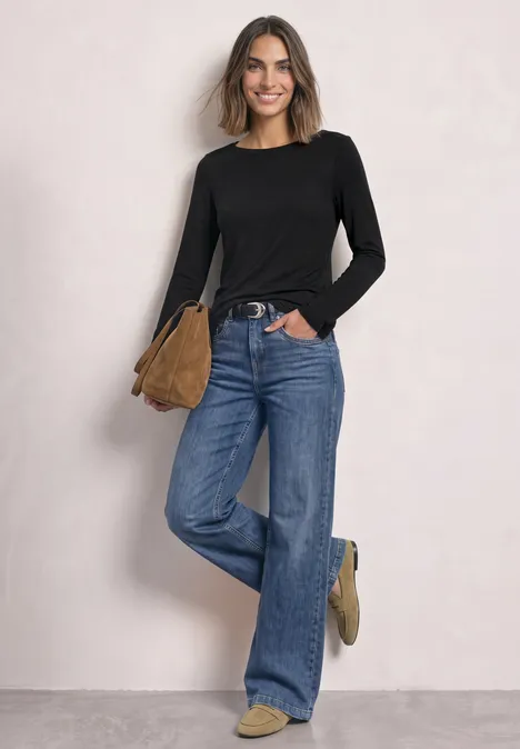 Basic Shirt mit U-Boot-Ausschnitt Black Eine lächelnde Frau in schwarzem Oberteil, weiter Jeans und brauner Tasche vor heller Wand.