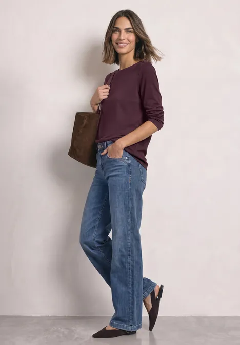 Vrouw in casual look met wijde jeans, bordeaux trui en een bruine tas.