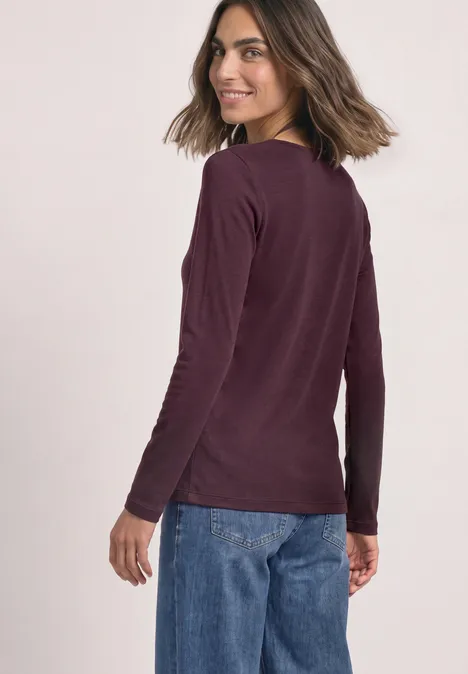 Een vrouw draagt een bordeauxrood shirt met lange mouwen en een blauwe spijkerbroek, kijkt over haar schouder.