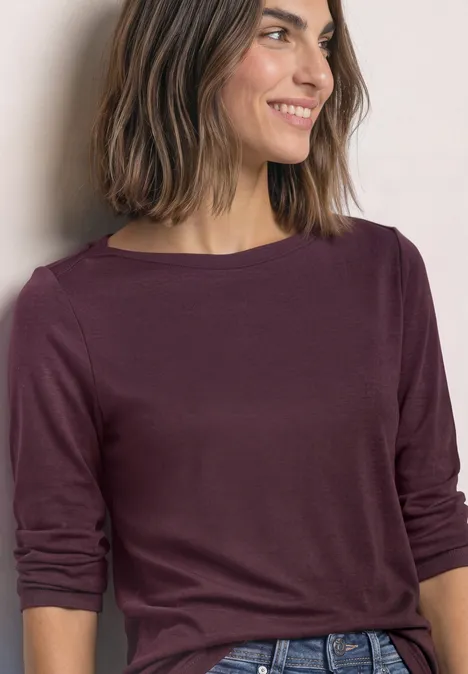 Vrouw met halflang haar in een bordeauxrode top met lange mouwen, glimlacht opzij.