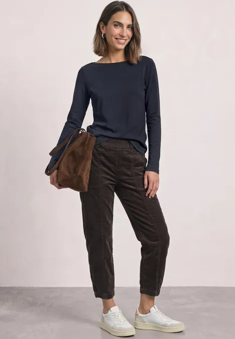 Femme souriante portant un pull bleu marine, pantalon marron, baskets blanches et sac assorti.