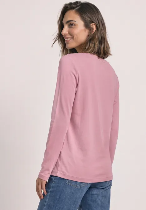 Eine Frau trägt ein rosafarbenes Langarmshirt und blaue Jeans, blickt lächelnd über die Schulter.