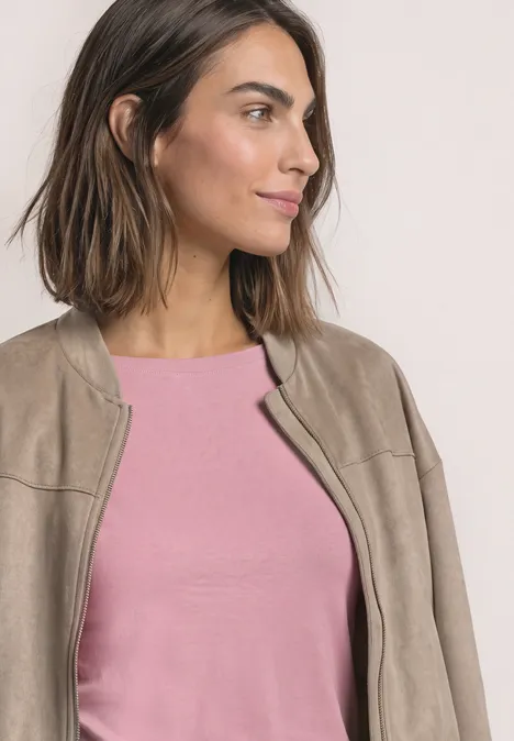 Eine lächelnde Frau mit schulterlangem Haar trägt ein rosafarbenes Oberteil und eine beige Jacke, Blick zur Seite.
