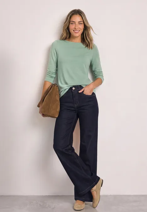 Basic Shirt mit U-Boot-Ausschnitt eucalyptus green Eine lächelnde Frau trägt eine mintgrüne Bluse, weite Jeans und flache Schuhe, hält eine braune Tasche.