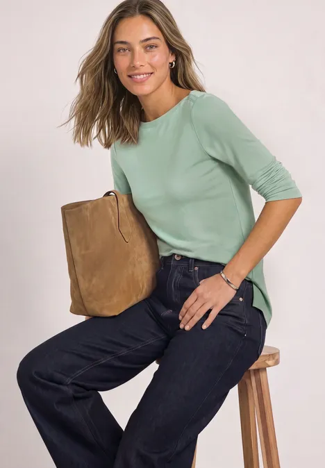 Basic Shirt mit U-Boot-Ausschnitt eucalyptus green Frau sitzt auf Hocker, trägt hellgrünes 3/4-Arm-Shirt, weite Jeans und hält braune Wildledertasche.