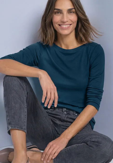 Haut basic a col bateau blue petrol Frau in langärmeligem, dunkelblauem Oberteil und Jeans sitzt lächelnd mit einem angewinkelten Bein.