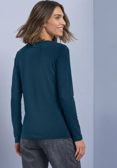 Haut basic a col bateau blue petrol Frau von hinten in dunkelgrünem Langarmshirt und Jeans vor hellgrauem Hintergrund.