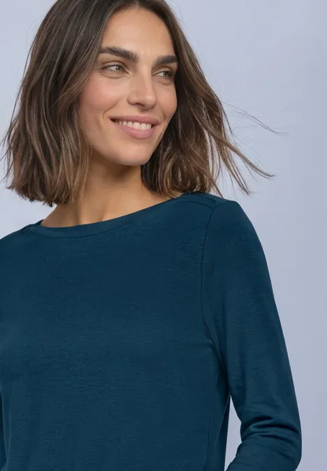 Haut basic a col bateau blue petrol Frau mit mittellangen braunen Haaren trägt ein blaugrünes Langarmshirt, leicht lächelnd vor hellem Hintergrund.