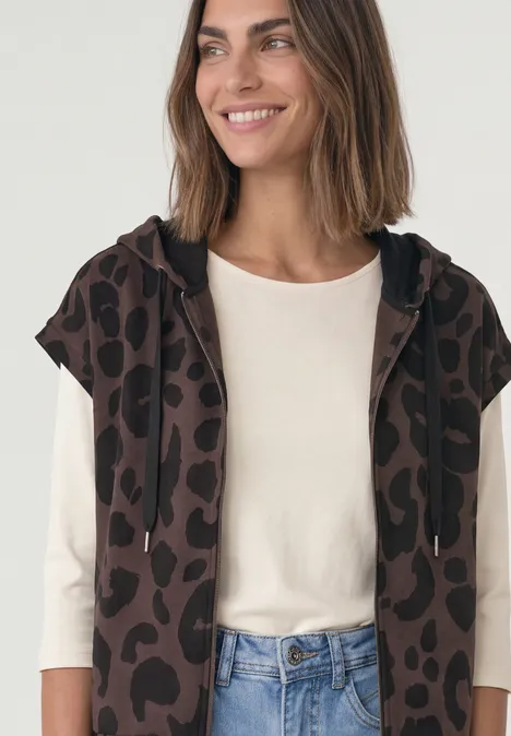 Frau in cremefarbenem Langarmshirt, offener ärmelloser Kapuzenpullover mit Leopardenmuster und Jeans.