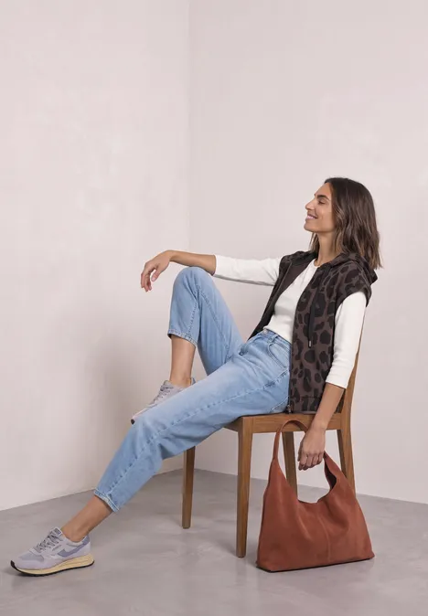 Weste mit Kapuze und Print molten earth Frau sitzt entspannt auf Stuhl, trägt weißes Shirt, Jeans, ärmellose Weste, Sneaker und Tasche.