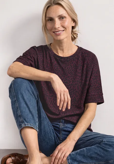 Eine lächelnde Frau sitzt entspannt in Jeans und einem gemusterten T-Shirt vor hellem Hintergrund.