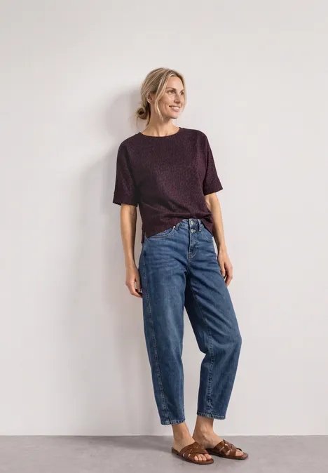 Eine lächelnde Frau lehnt an einer hellen Wand, trägt lila Shirt, hochtaillierte Jeans mit Umschlag und braune Sandalen.
