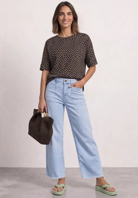 Frau in gepunktetem T-Shirt, hellblauen, weiter geschnittenen Jeans und grünen Sandalen mit brauner Tasche.