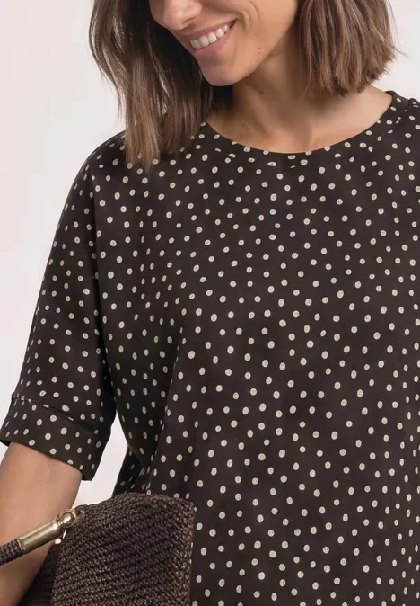 Frau trägt schwarze Bluse mit weißen Punkten und hält eine braune gewebte Tasche.