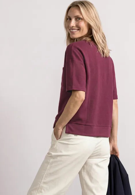 Femme portant un sweat bordeaux à manches courtes et un pantalon clair, souriante en se retournant.