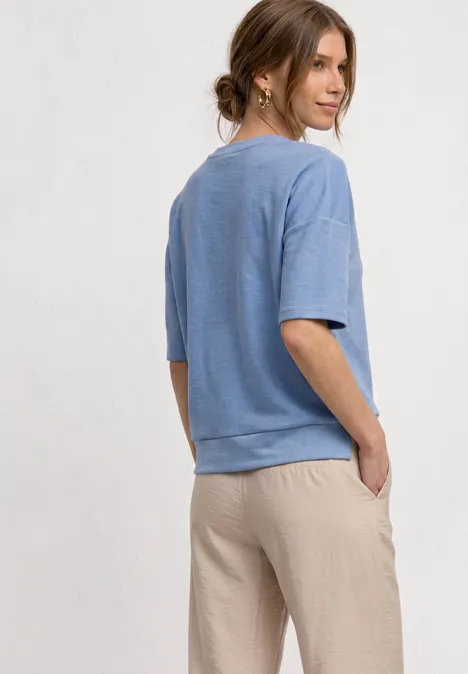 Shirt met ronde hals en textuur northern blue Vrouw in lichtblauwe korte mouw trui en beige broek kijkt over haar schouder tegen neutrale achtergrond.