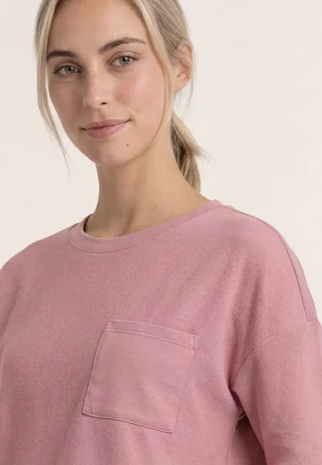 Shirt met ronde hals en structuur vintage blush Een jonge vrouw met blond haar in een paardenstaart draagt een roze shirt met borstzak, zachte blik en neutrale achtergrond.