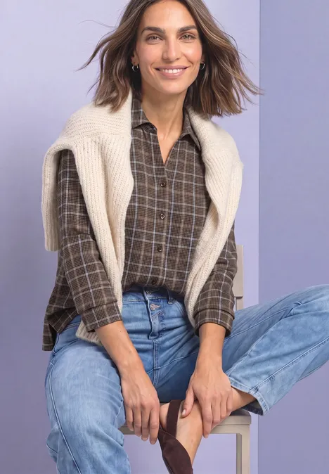 Eine Frau sitzt lächelnd, trägt ein kariertes Hemd, hellen Pullover über den Schultern und blaue Jeans.
