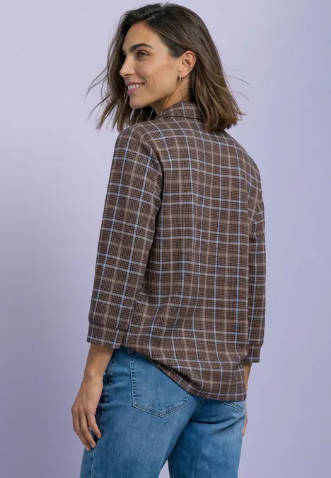 Frau mit schulterlangen Haaren trägt kariertes Hemd und Jeans, steht seitlich und lächelt.