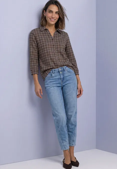 Frau in braun-karierter Bluse und hellblauen Jeans vor lila Hintergrund.