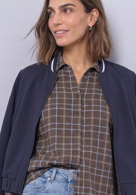 Eine lächelnde Frau mit brünettem Haar trägt eine karierte Hemdbluse und eine dunkelblaue Bomberjacke.