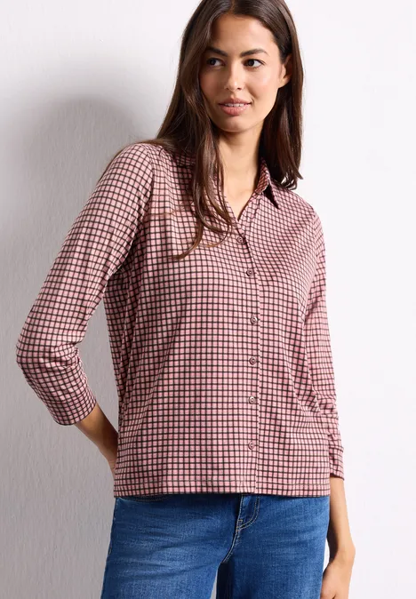 Eine Frau trägt eine rot-weiß karierte Bluse und blaue Jeans vor einer weißen Wand.