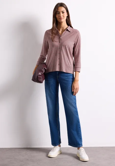 Frau in rosa Hemd, blauen Jeans und weißen Sneakern vor weißem Hintergrund.