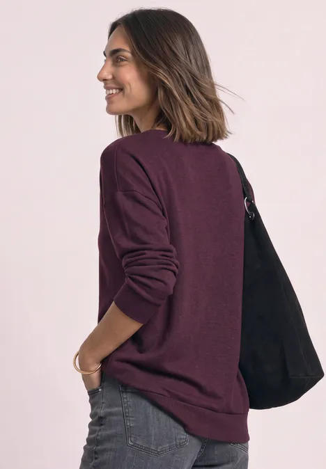 Frau in seitlicher Ansicht mit weinrotem Pullover, Jeans und schwarzer Tasche, lächelnd.