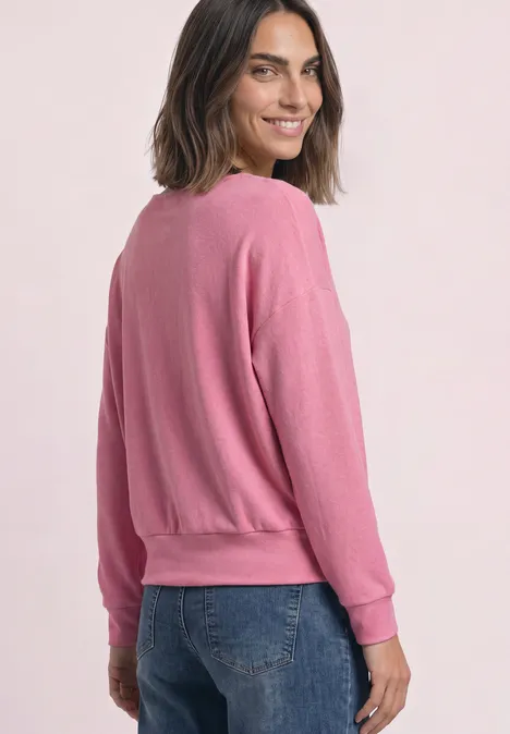 T-shirt à manches longues avec ourlet élastiqué warm blush mel. Femme aux cheveux mi-longs porte un pull rose et un jean bleu, sourit en regardant par-dessus son épaule.