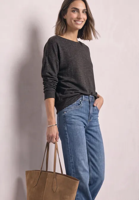 Frau in blauem Jeans, dunklem Pullover mit großer brauner Tasche, lächelnd vor hellgrauem Hintergrund