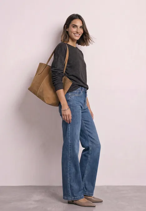Eine lächelnde Frau mit schwarzem Pullover, weitem Jeans und brauner Tasche vor hellem Hintergrund.
