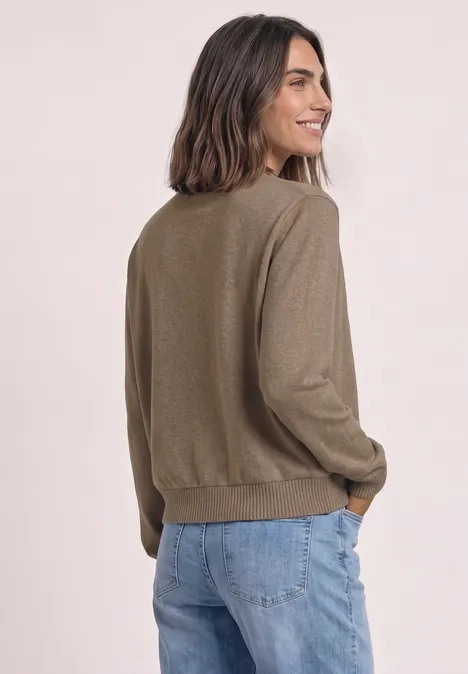 Eine Frau in blauen Jeans und olivgrünem Pullover blickt über die Schulter und lächelt.