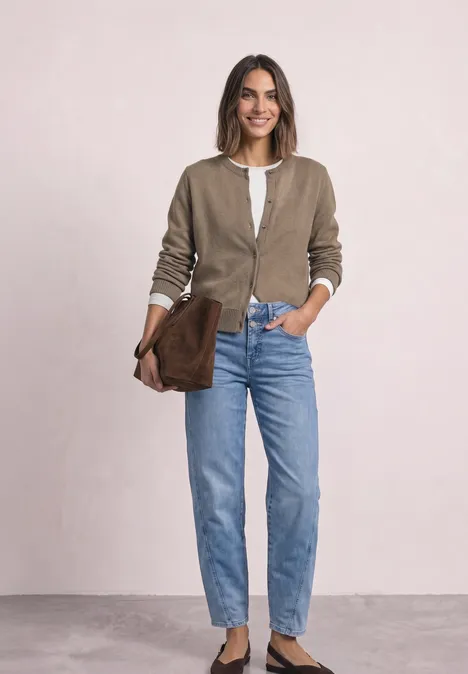 Frau lächelt, trägt beige Cardigan, weißes Top, blaue Jeans und hält eine braune Clutch vor hellem Hintergrund.