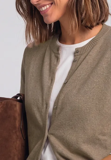Frau mit schulterlangem braunem Haar trägt einen khakifarbenen Cardigan über weißem T-Shirt und hält eine Tasche.