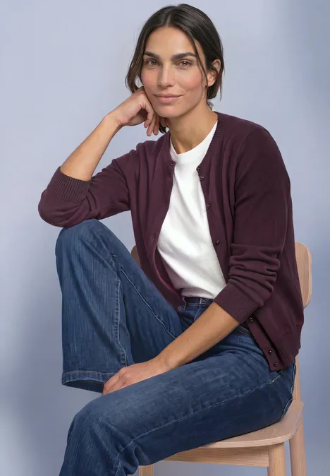 Femme en cardigan violet, t-shirt blanc et jean, assise décontractée sur un tabouret, appuie sa tête sur la main.