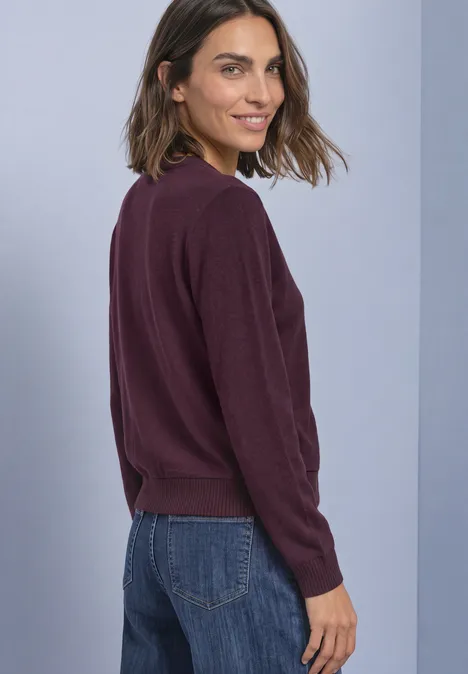 Femme en pull violet foncé et jean, souriante, tourne la tête par-dessus son épaule.