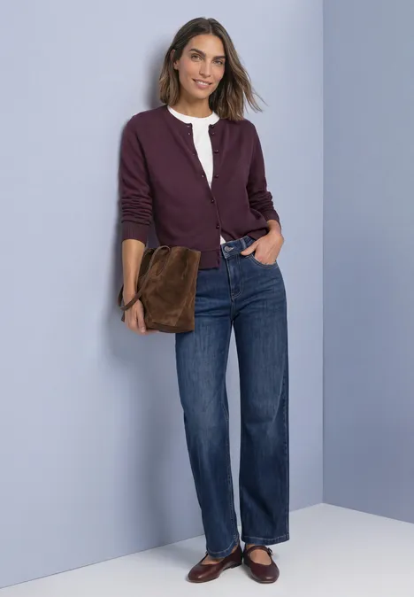 Femme souriante en blouse blanche, gilet violet, jean droit et mocassins marron, tenant une pochette marron.
