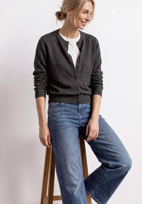 Frau lehnt lächelnd an einem Hocker, trägt grauen Strickcardigan, weißes Shirt und weite blaue Jeans.