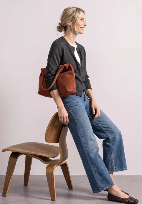 Frau in grauer Strickjacke, weißer Bluse und weiten Jeans, trägt braune Ledertasche und steht neben einem Holzstuhl.