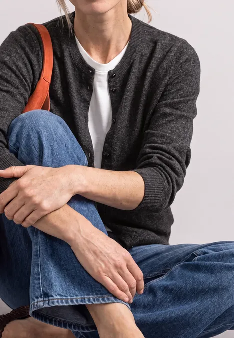 Frau in Jeans, weißem T-Shirt und dunkelgrauem Cardigan sitzt, Hand auf dem Knie, orangefarbener Taschenriemen.