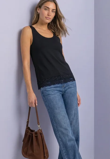 Frau mit schwarzem Top, Jeans und brauner Handtasche vor neutralem Hintergrund.