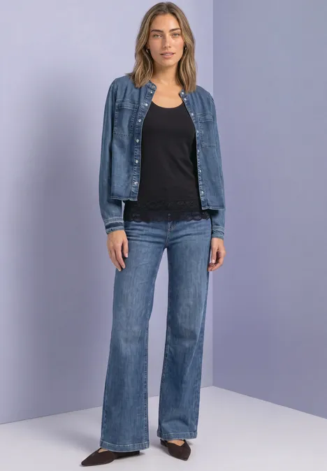 Eine Frau in schwarzer Bluse, Jeansjacke und ausgestellten Jeans vor neutralem Hintergrund.