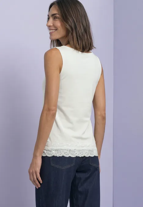 Frau in weißem, ärmellosem Top mit Spitzensaum und dunkler Jeans blickt lächelnd vor lila Hintergrund.