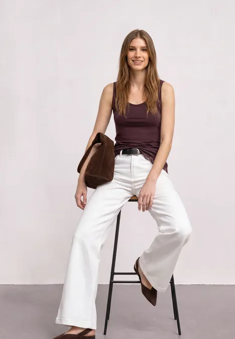 Frau mit braunem Top und weißer Schlaghose sitzt lächelnd auf einem Hocker und hält eine braune Tasche.