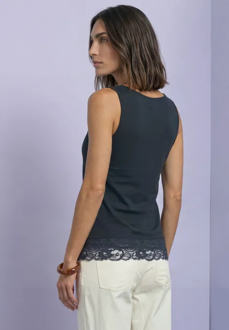 Dame trägt ärmelloses schwarzes Top mit Spitzenbesatz und helle Hose vor hellem Hintergrund.