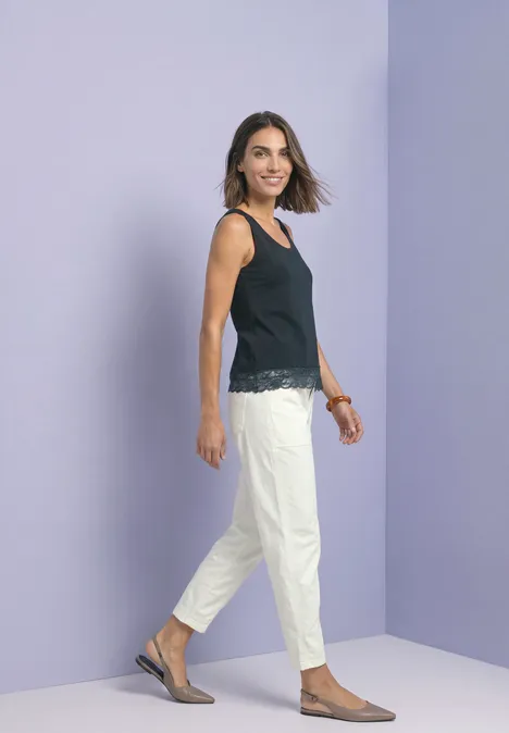 Eine Frau lächelt in schwarzem Top und weißer Hose vor lila Wand.
