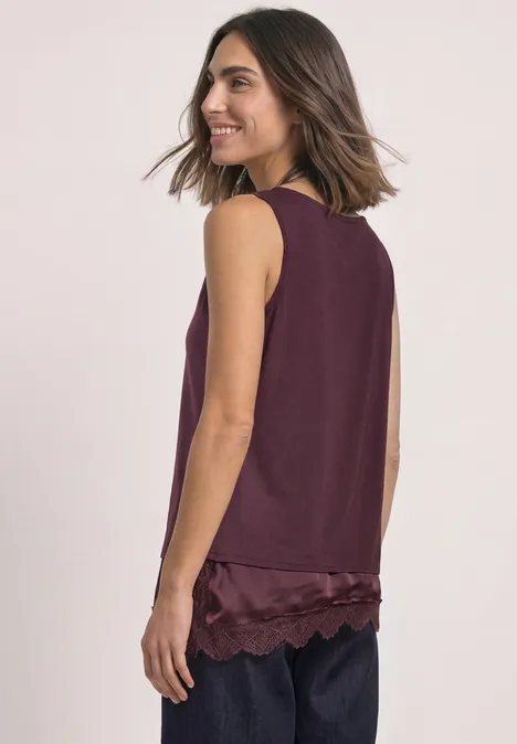 Vrouw in zijaanzicht met een mouwloos bordeauxrood topje met kanten zoom en een donkere broek.