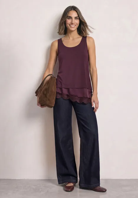 Vrouw in paarse, mouwloze top met laagjes, wijde donkere broek, bruine sandalen en schoudertas.