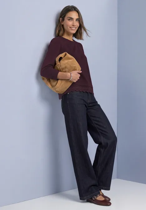 Frau in weinrotem Pullover, weiten Jeans und braunen Loafers mit gelber Tasche vor hellem Hintergrund.
