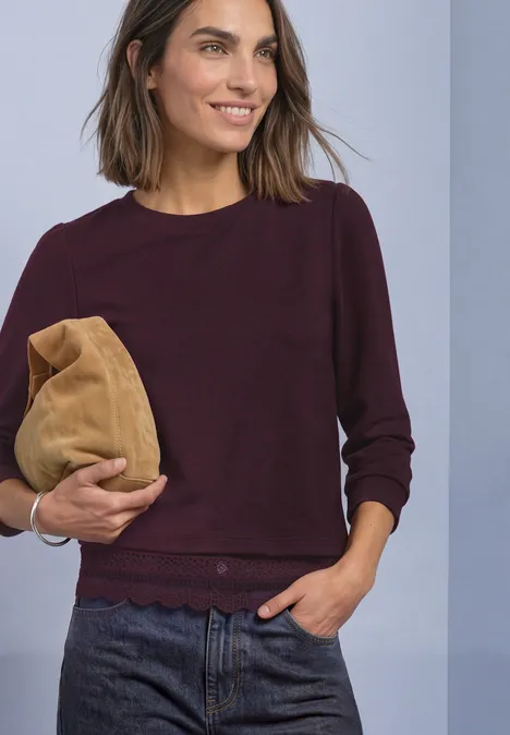 Frau trägt dunkelrotes Sweatshirt und hält eine beige Clutch, lächelt dabei natürlich.